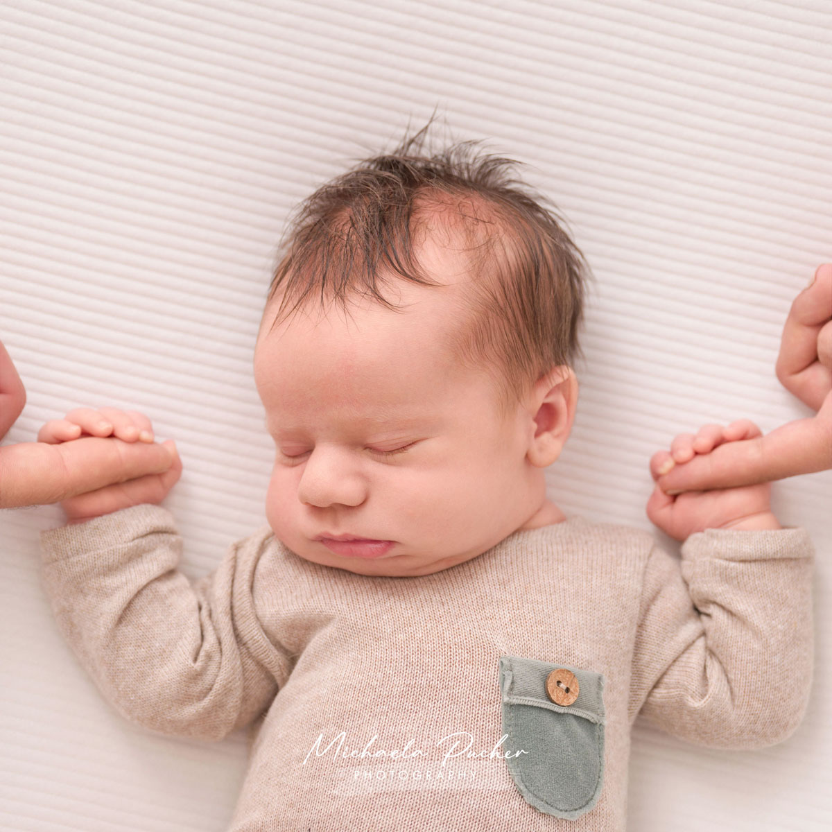 Schlafendes Neugeborenes hält links und rechts die Finger seiner Geschwister, während es in einem beigefarbenen Strampler auf einer weißen Unterlage liegt.